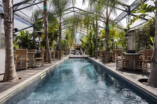 Indoor glass conservatory with a narrow pool, wooden deck, palm trees, wicker chairs, and large E&M letters at the far end.