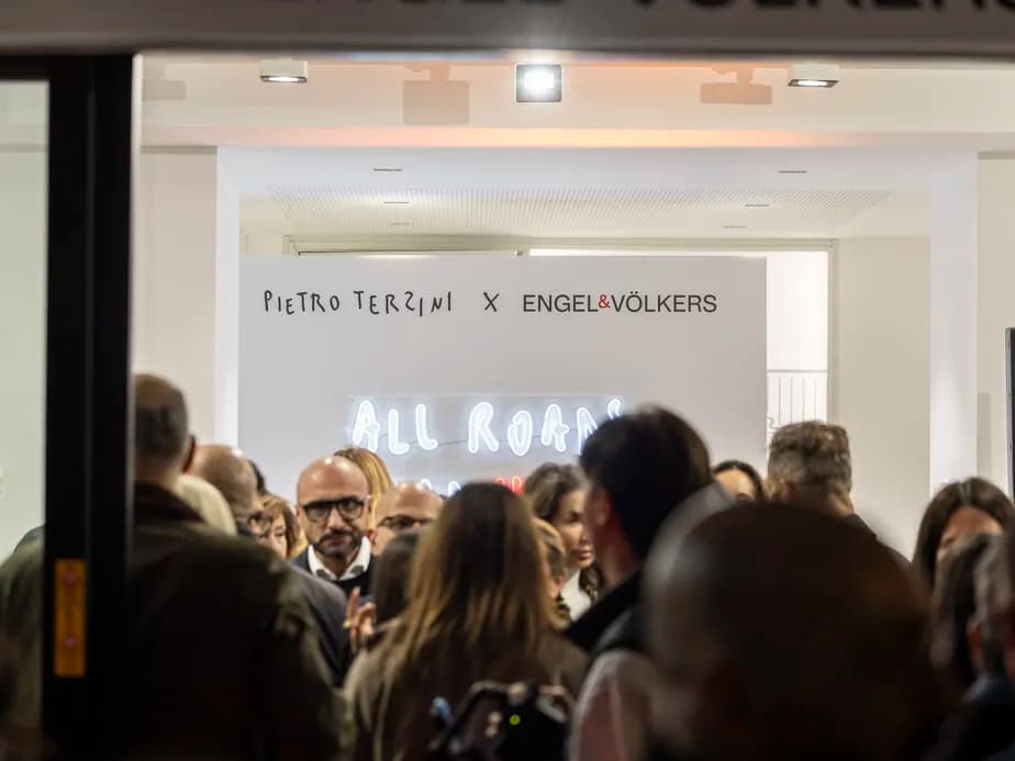 Crowded gallery entrance featuring a Pietro Terzini x Engel & Völkers sign, white walls, and a glowing neon sign among many people.