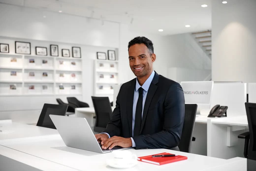 Smiling man in a dark suit sits at a white desk with a laptop in a bright, modern office; shelves with framed photos behind him.