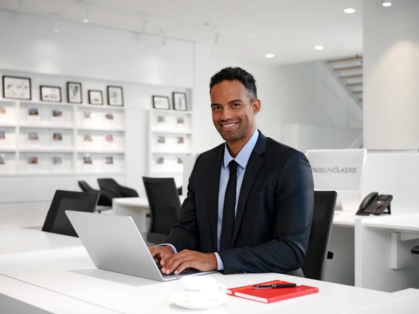 Smiling man in a dark suit sits at a white desk with a laptop in a bright, modern office; shelves with framed photos behind him.