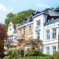 Eine Reihe charmanter, farbenfroher Häuser im europäischen Stil mit Balkonen, umgeben von Grün und Bäumen unter einem klaren blauen Himmel.