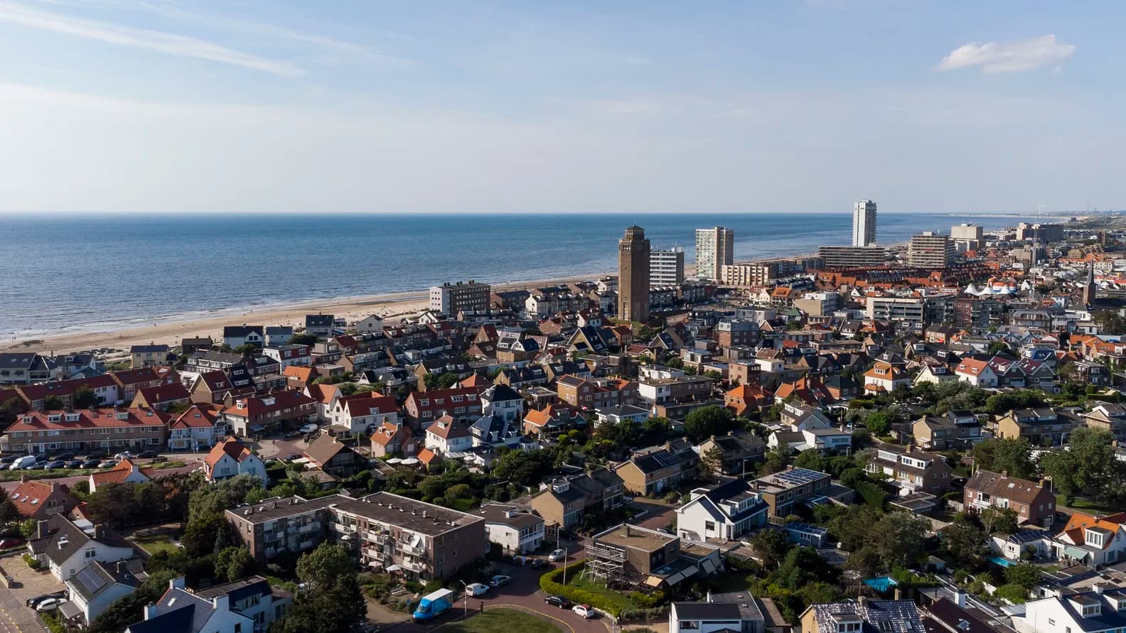 Aerial view of a coastal town with red-roofed houses, a sandy beach, and the blue sea under a clear sky.