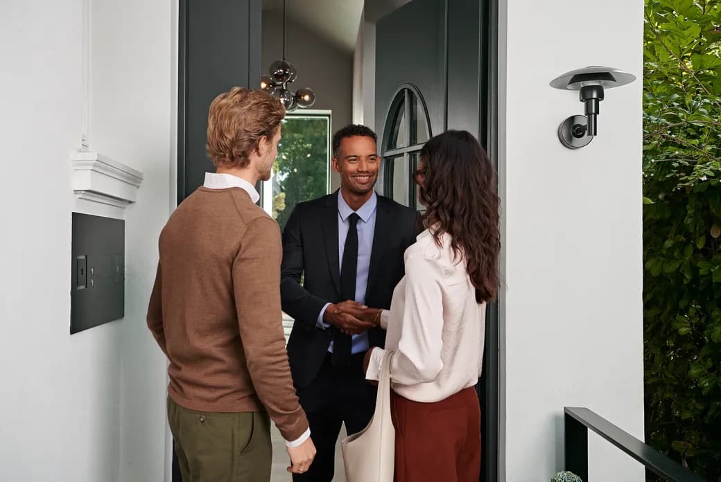 A real estate agent in a suit greets a couple at the doorway of a modern white house, with greenery outside.