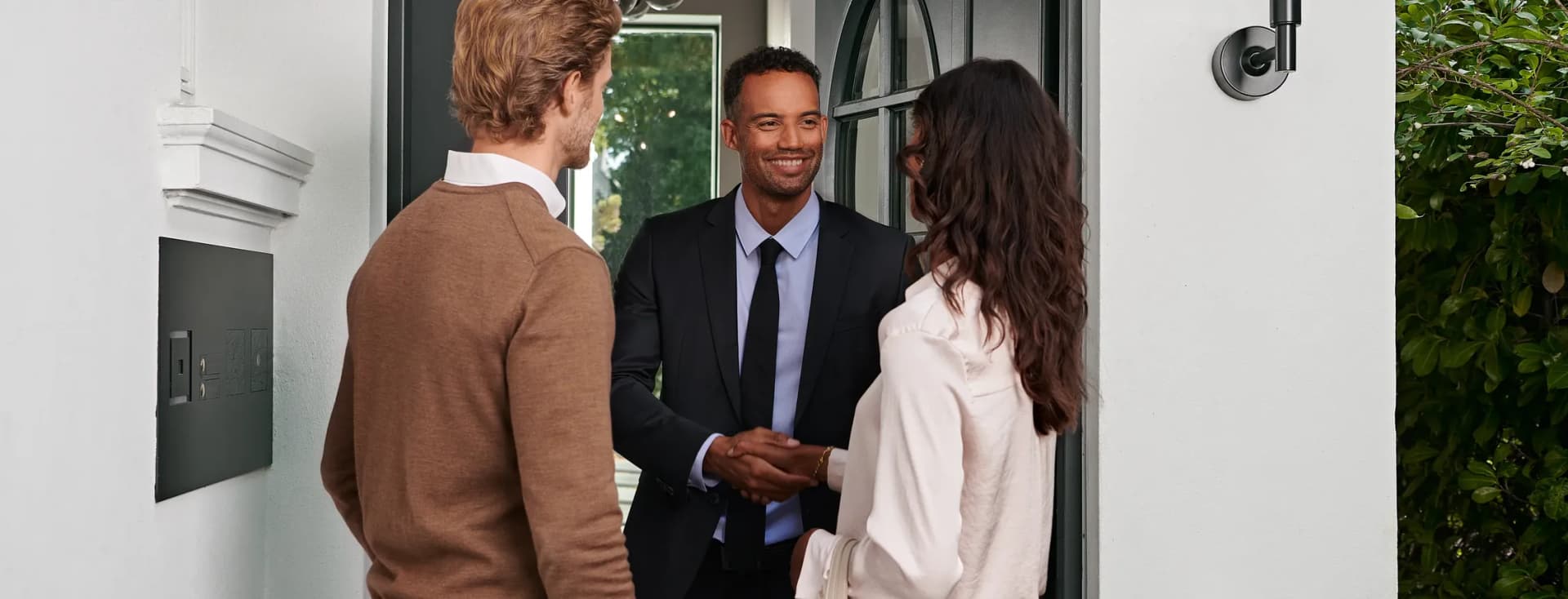 A real estate agent in a suit greets a couple at the doorway of a modern white house, with greenery outside.