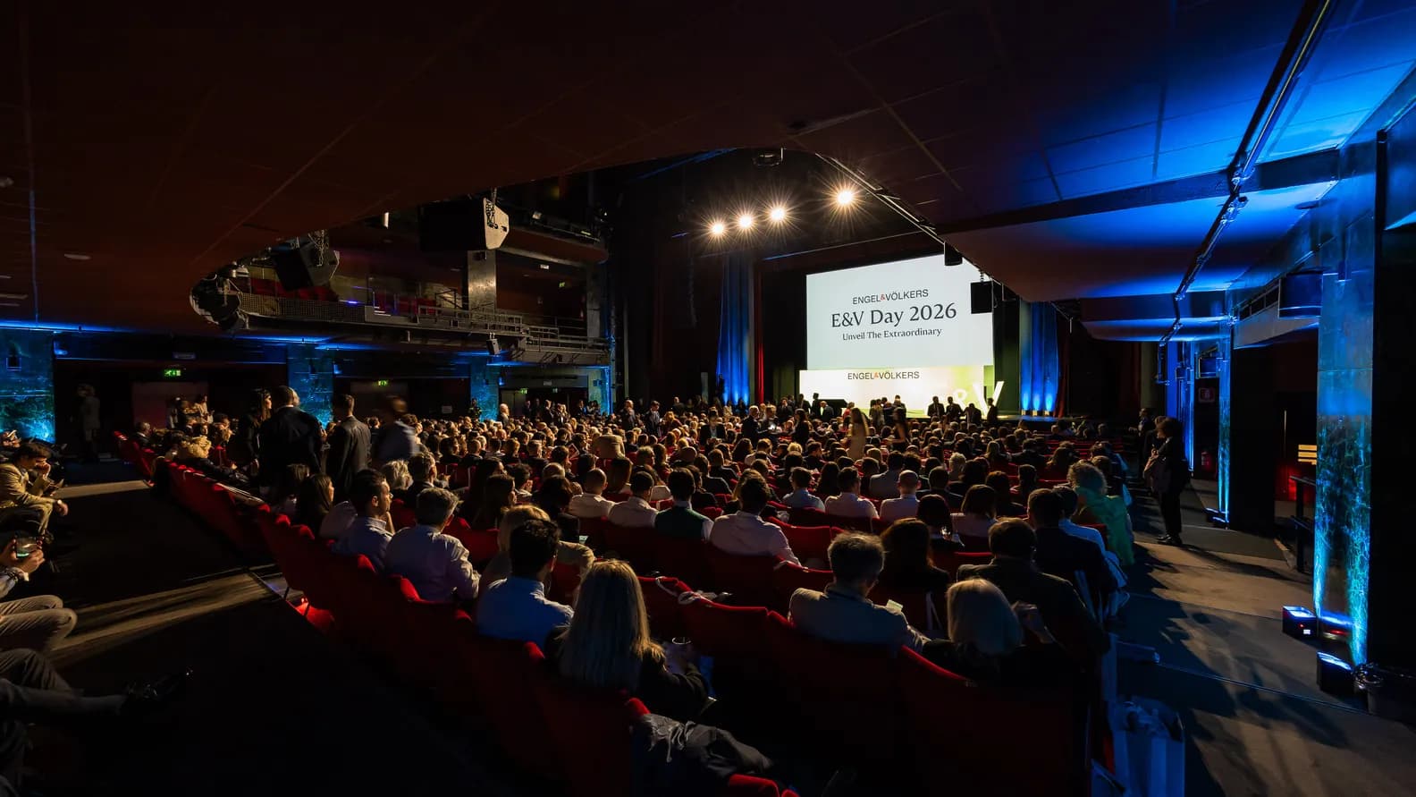 A crowded conference hall with rows of red seats, attendees facing a stage and a large screen reading “E&V Day 2026” under bright spotlights.