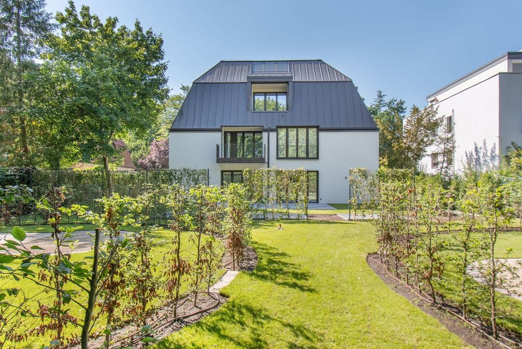 Modern white house with a dark metal mansard roof and large windows, set in a landscaped garden with trees and a low hedge.