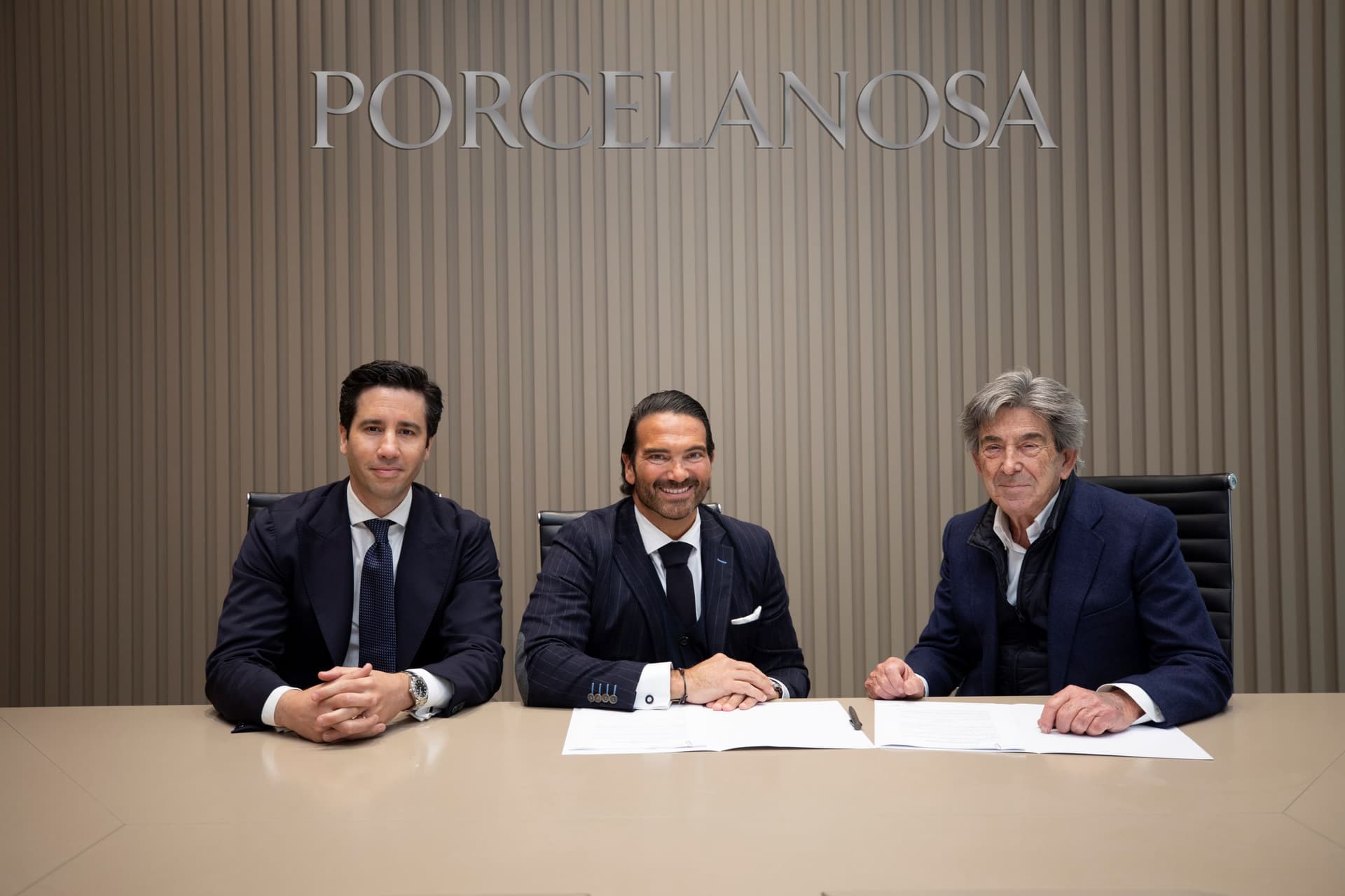 Tres homes en vestits s'asseuen a una taula amb documents signats davant d'un rètol de "Porcelanosa".
