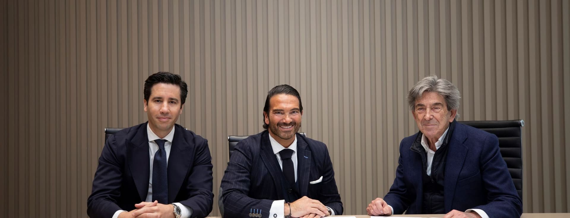 Tres homes en vestits s'asseuen a una taula amb documents signats davant d'un rètol de "Porcelanosa".