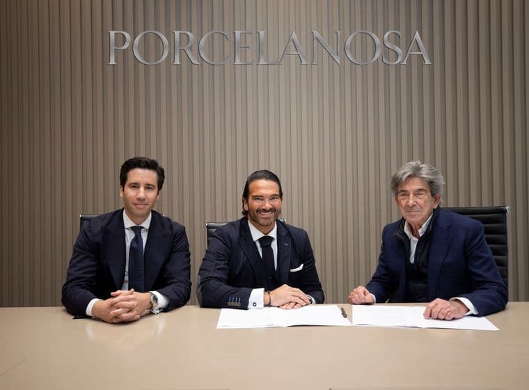 Tres homes en vestits s'asseuen a una taula amb documents signats davant d'un rètol de "Porcelanosa".
