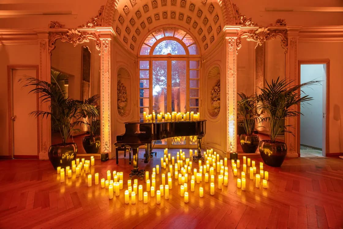 Ornate Rococo room with a black grand piano, rows of candles, tall potted plants, framed by an arched window.