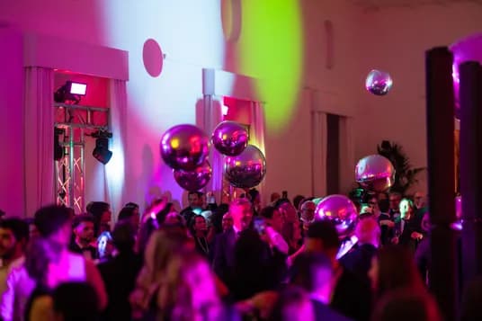 A lively indoor party scene bathed in pink, purple, and yellow lights; a packed crowd beneath floating reflective balloons.