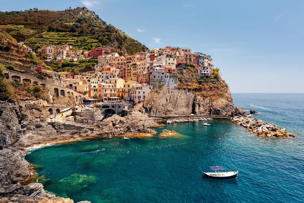 Colorful cliffside village with pastel buildings on terraced hills overlooking turquoise sea and small boats.