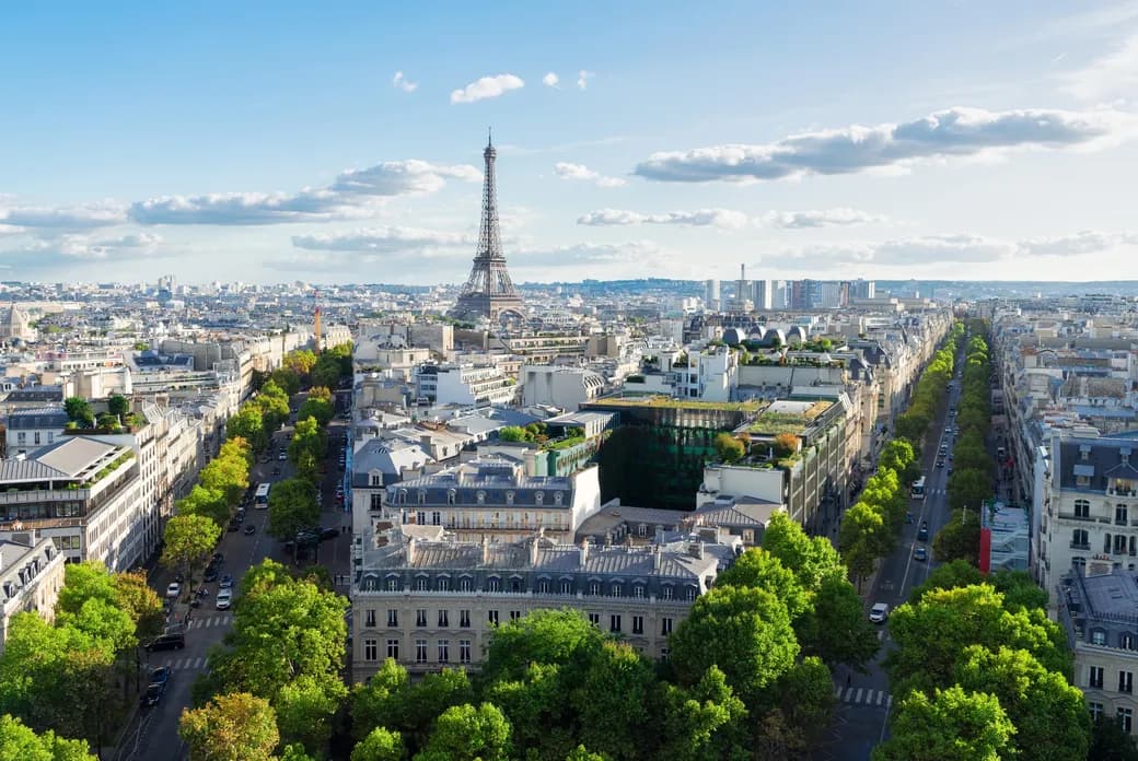 Aerial view of Paris with the Eiffel Tower in the distance, surrounded by historic buildings and tree-lined streets under a partly cloudy sky.
