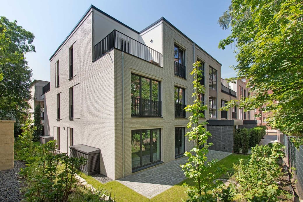 Contemporary light‑brick apartment building with black metal balcony rails, large windows and surrounding trees.