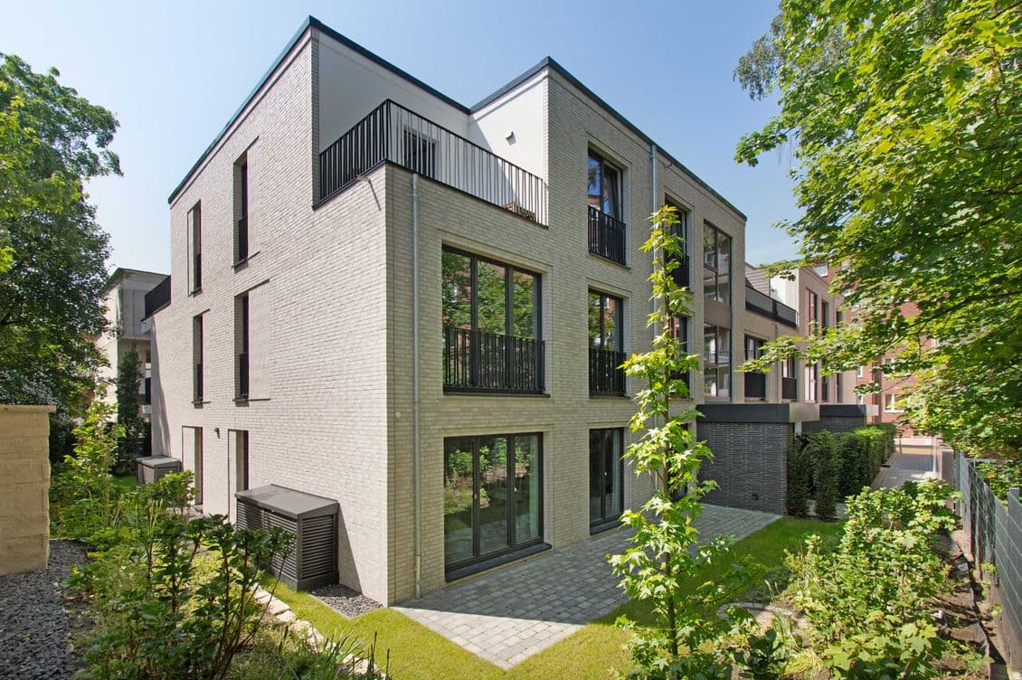 Modernes Apartmenthaus aus hellem Backstein mit schwarzen Metallgeländern an den Balkonen, großen Fenstern und Bäumen in der Umgebung.