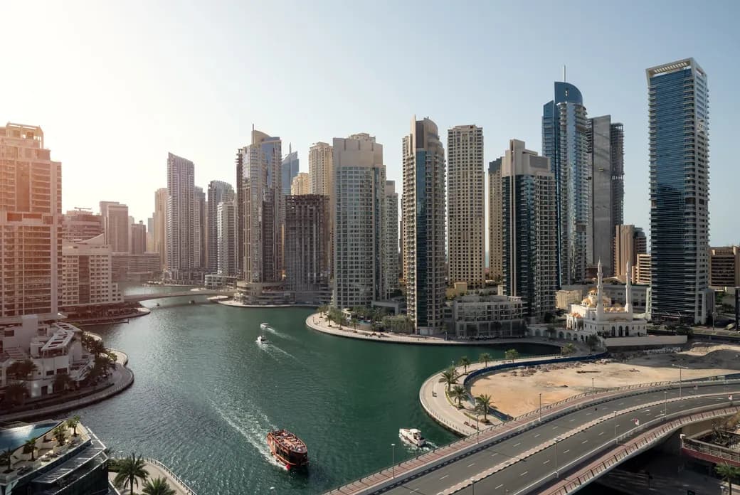 Sunlit city skyline of modern high-rises along a winding canal, with boats on the water and a curved highway in the foreground.