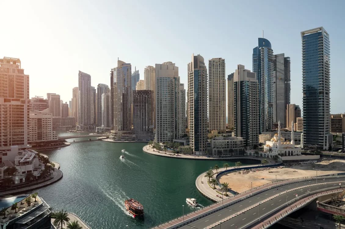 Sunlit city skyline of modern high-rises along a winding canal, with boats on the water and a curved highway in the foreground.