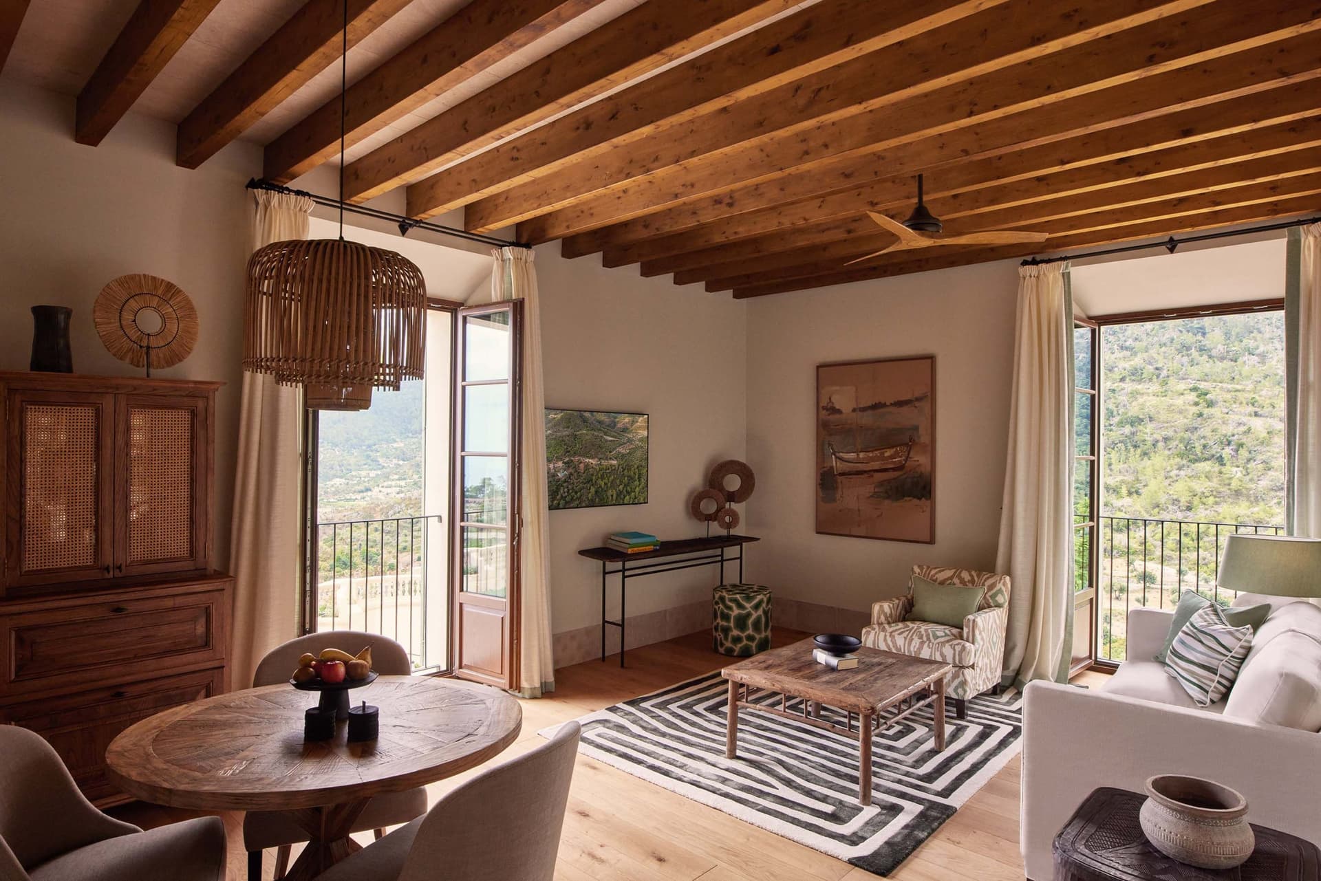 Helles Wohnzimmer mit Holzbalkendecke, offenem Balkon und neutralen Möbeln. Ein runder Holztisch und Stühle stehen in der Nähe eines dunklen Holzschranks.