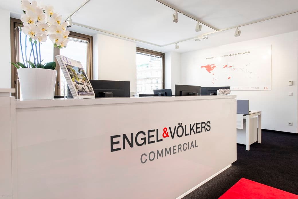 Modern office reception with glossy white desk, displaying "ENGEL & VÖLKERS COMMERCIAL" logo, orchid plant, and marketing materials.