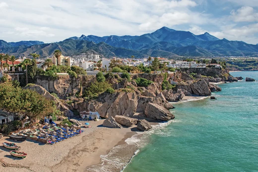 Coastal town perched on rocky cliffs with white buildings and palm trees, turquoise sea, and a sandy beach with blue umbrellas, mountains in the distance.