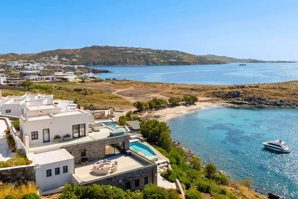 White cubic houses with flat roofs line a hillside along a blue bay; calm water, rocky coastline, and a small yacht anchored nearby.