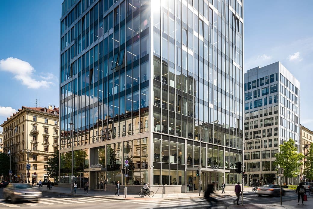 Modern glass office building on a busy city street corner, with pedestrians and cyclists under a clear blue sky.
