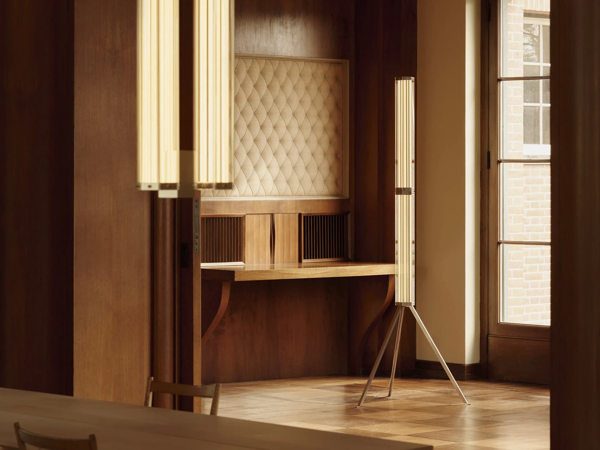 Warm wood-paneled room with a padded quilted wall panel, two tall vertical light fixtures, a wooden desk, and a window showing brick exterior.