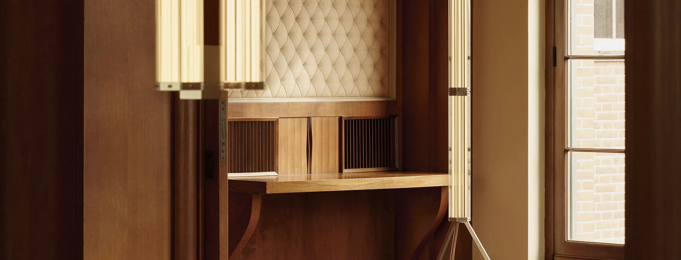 Warm wood-paneled room with a padded quilted wall panel, two tall vertical light fixtures, a wooden desk, and a window showing brick exterior.