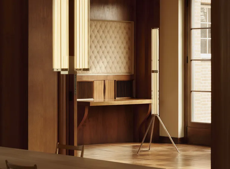 Warm wood-paneled room with a padded quilted wall panel, two tall vertical light fixtures, a wooden desk, and a window showing brick exterior.