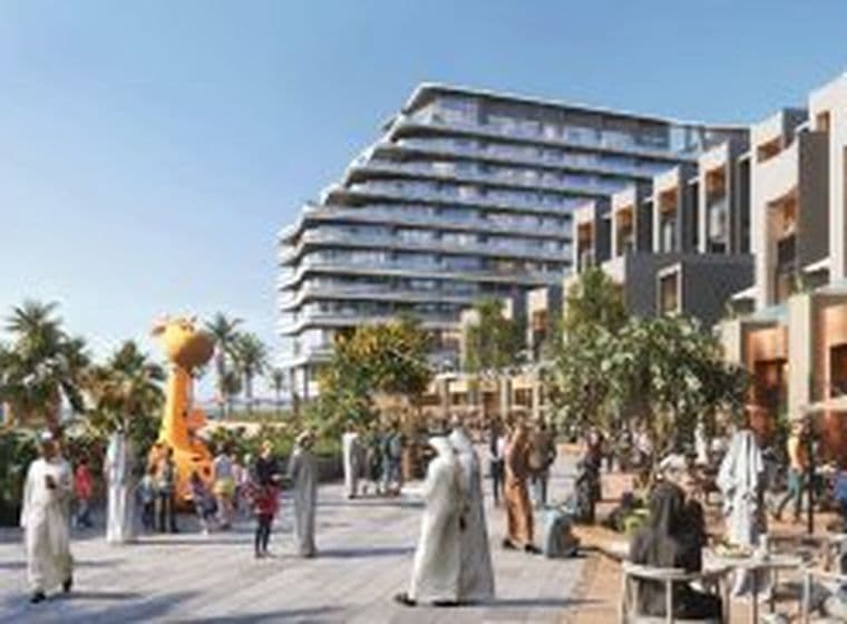 Outdoor plaza by a modern seaside building with people strolling and socializing, featuring palm trees and a giant yellow giraffe sculpture.