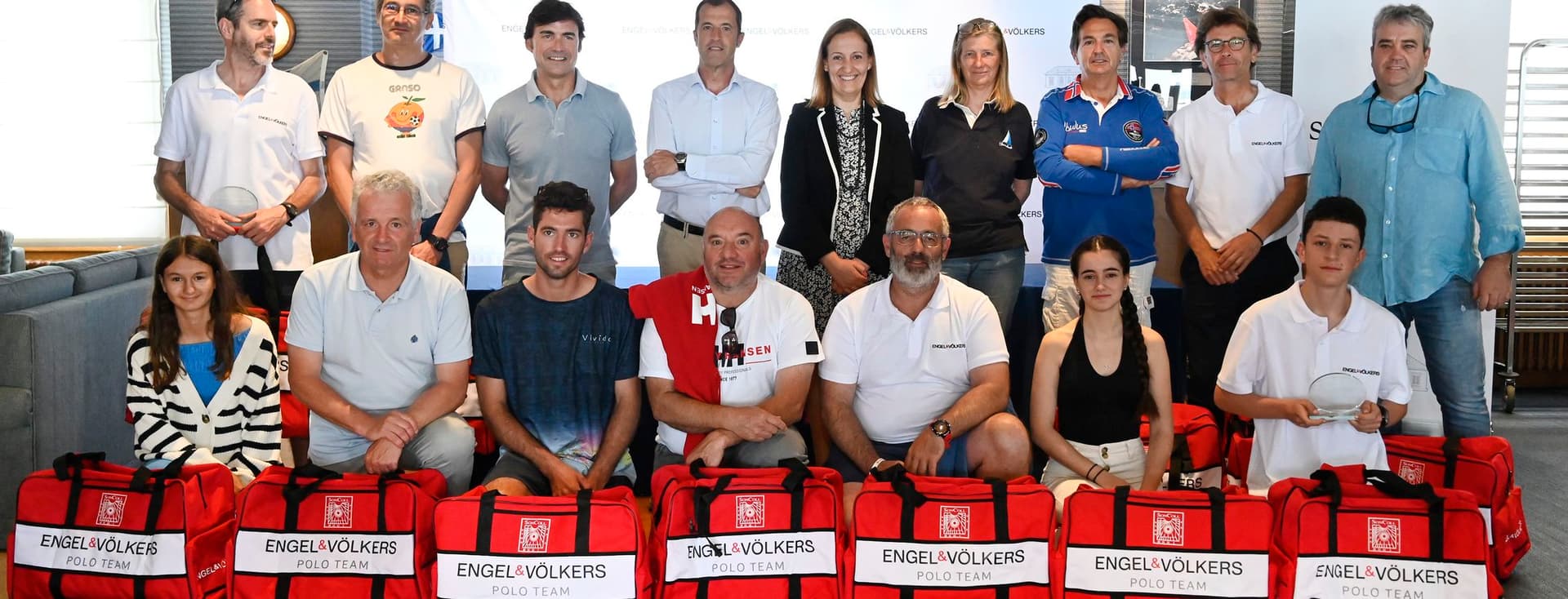 Un grup de 17 persones posen amb bosses vermelles de l'equip de polo d'Engel & Völkers. Alguns estan dempeus, alguns agenollats. Hi ha una bandera cubana al fons.