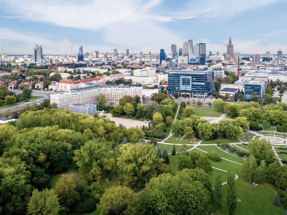 Lotniczy widok na panoramę miasta z nowoczesnymi wieżowcami, bujnym zielonym parkiem i rozproszonymi chmurami na błękitnym niebie.