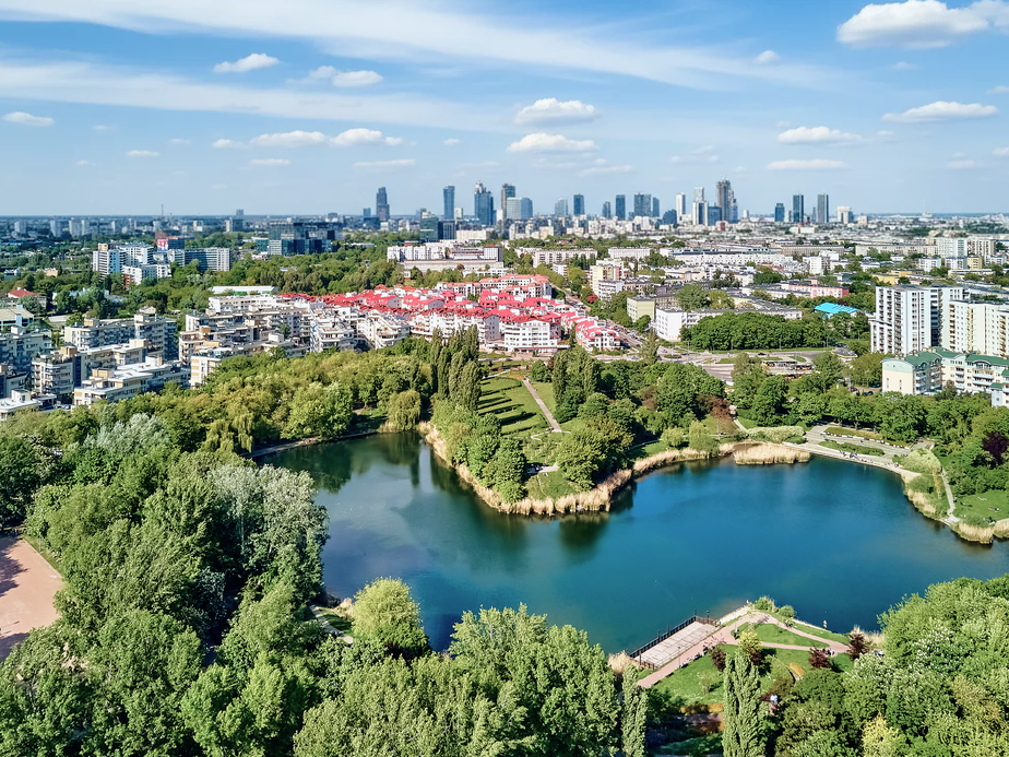 Aerial view of a cityscape with a green park, lake, and modern buildings under a partly cloudy sky.