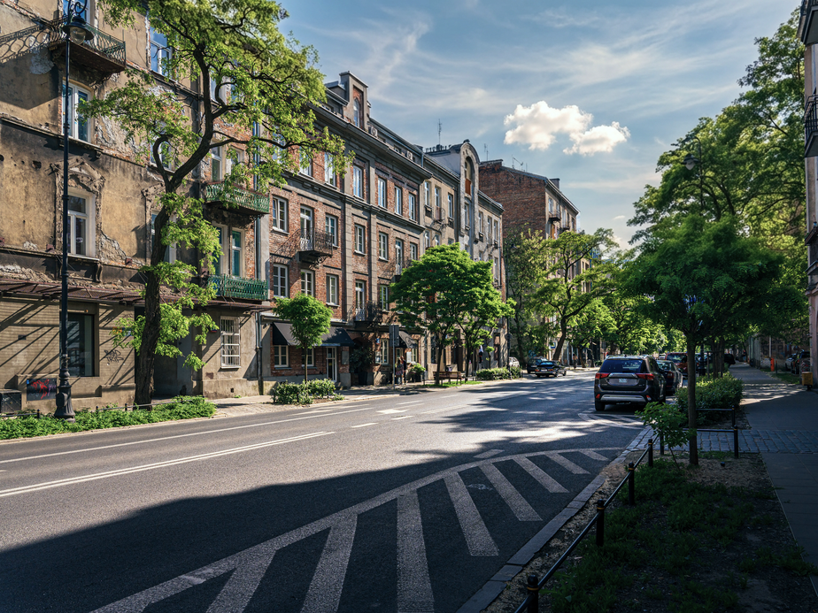 Sunny street with historic buildings, trees lining the sidewalk, parked cars, and a striped pedestrian crossing under a partly cloudy sky.