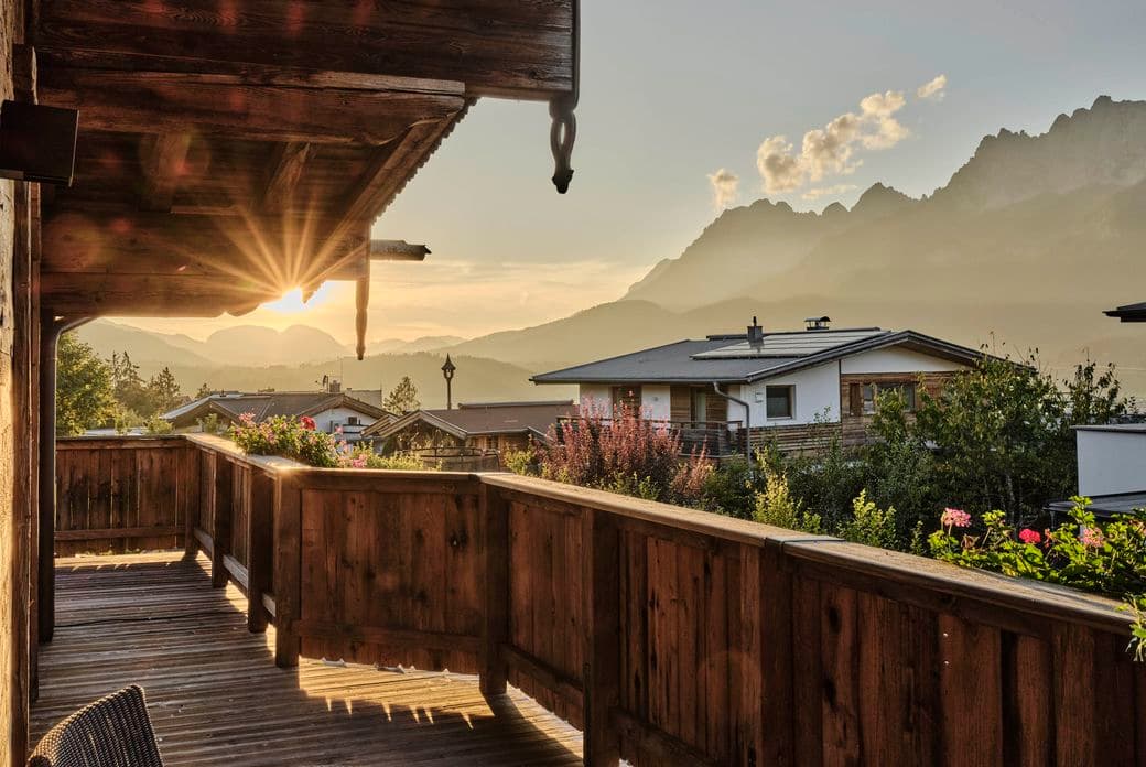 View from a wooden balcony with flowers, overlooking houses and mountains at sunset.