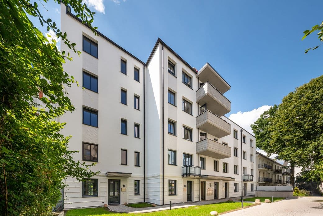 Modern white apartment building with balconies and dark-framed windows, surrounded by green trees under a clear blue sky.