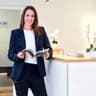 Smiling woman in a navy blazer and white shirt, holding a magazine, stands by a reception desk with orchids in a modern office setting.