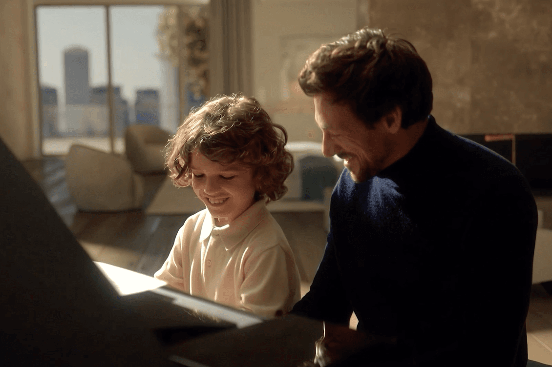 A man and a child sit together at a piano, smiling, in a sunlit room with large windows and modern furniture.
