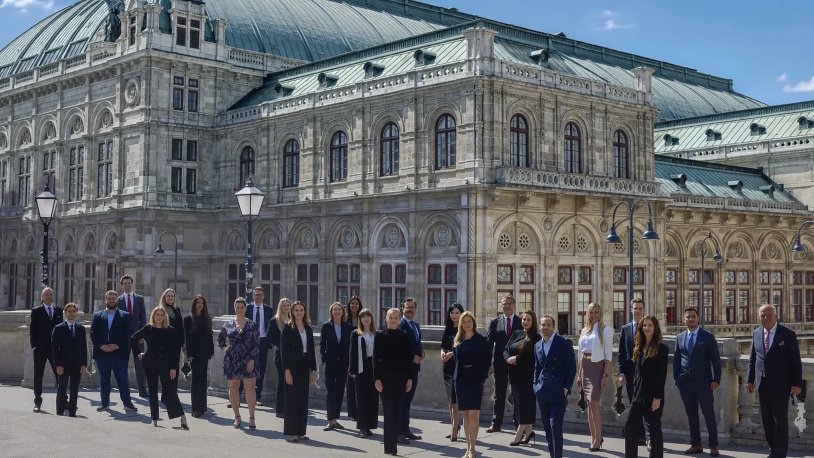 Eine Gruppe von Geschäftsleuten posierte vor einem prächtigen, reich verzierten Steingebäude mit einem grünen Kupferdach unter strahlend blauem Himmel.