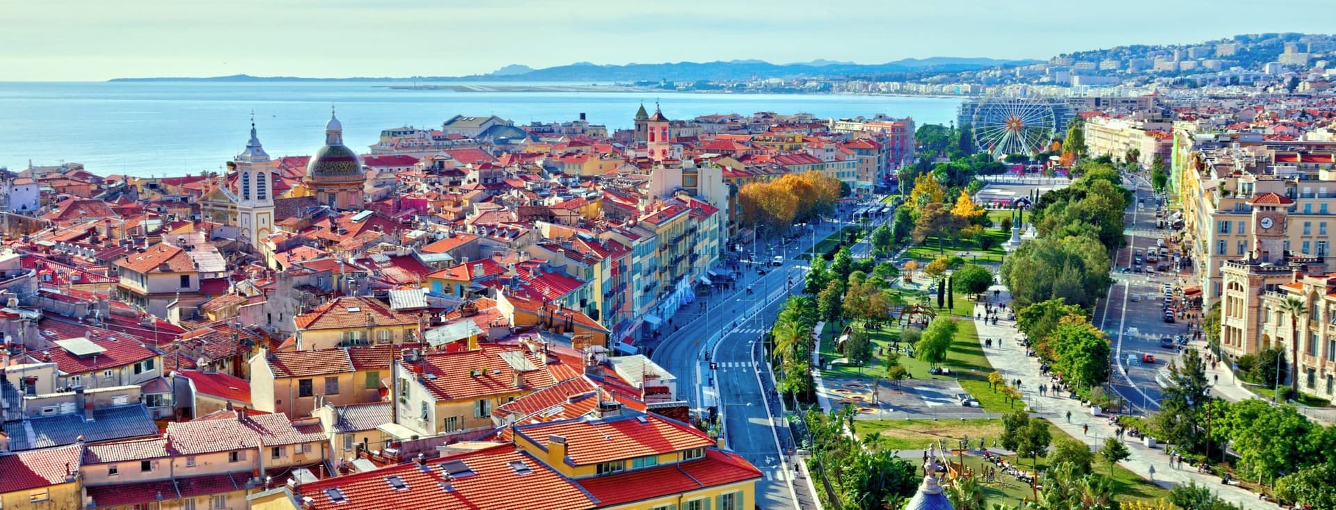Aerial view of Nice, France, featuring red-tiled roofs, a Ferris wheel, and the Mediterranean Sea under a blue sky.
