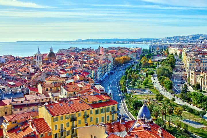 Aerial view of Nice, France, featuring red-tiled roofs, a Ferris wheel, and the Mediterranean Sea under a blue sky.