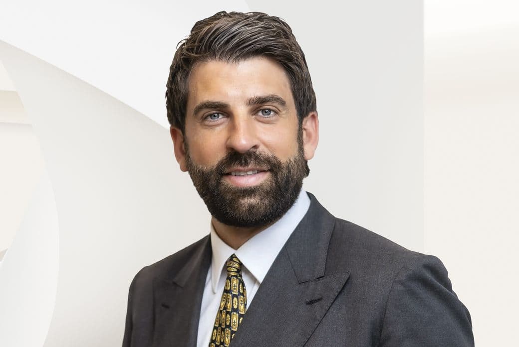 Smiling bearded man in dark suit and patterned tie, close-up headshot against a light geometric background.