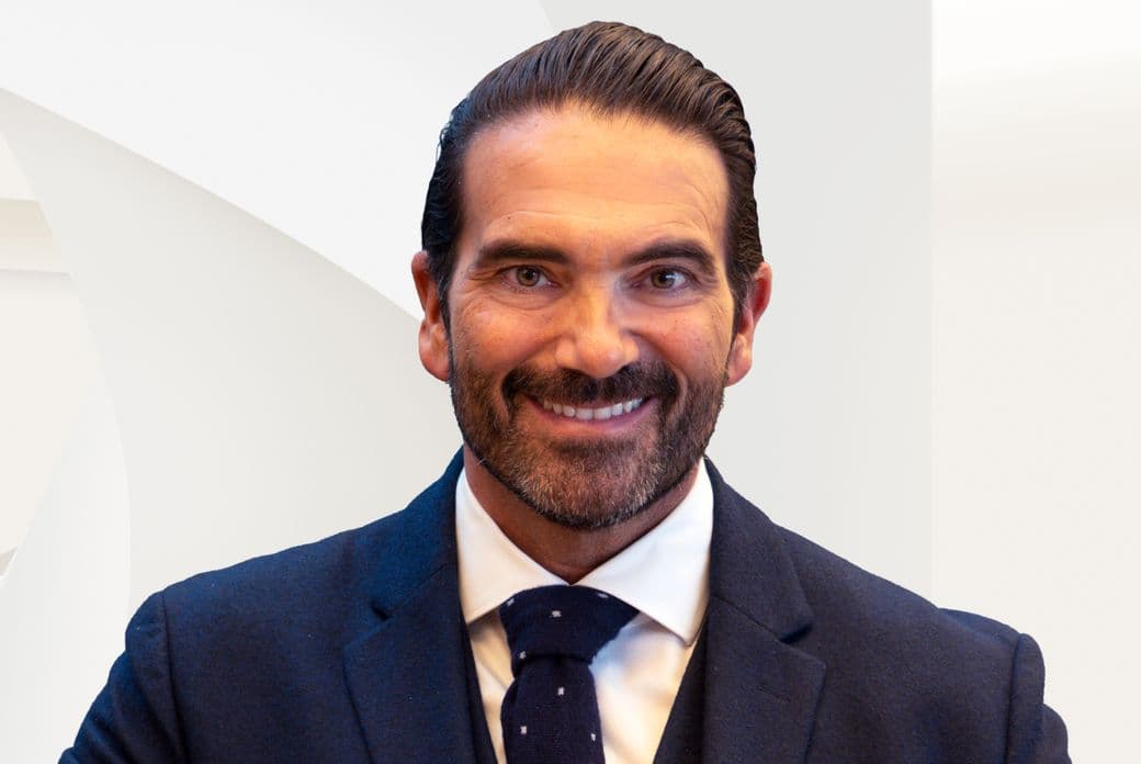 Close-up of a smiling bearded man wearing a navy suit, white shirt and patterned tie against a light geometric background.