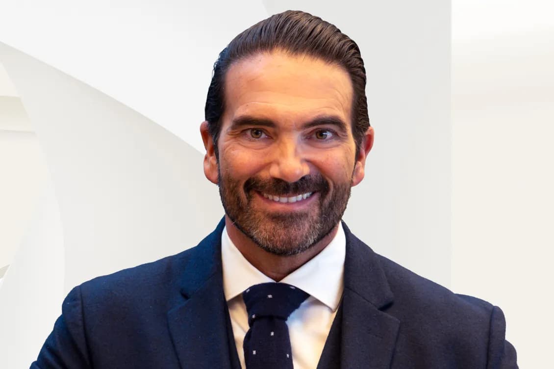 Close-up of a smiling bearded man wearing a navy suit, white shirt and patterned tie against a light geometric background.