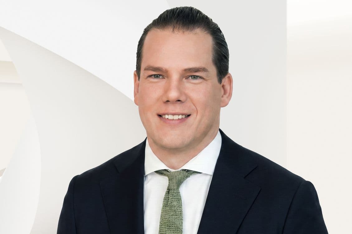 Smiling man in dark suit and green tie, close-up headshot against a light geometric background