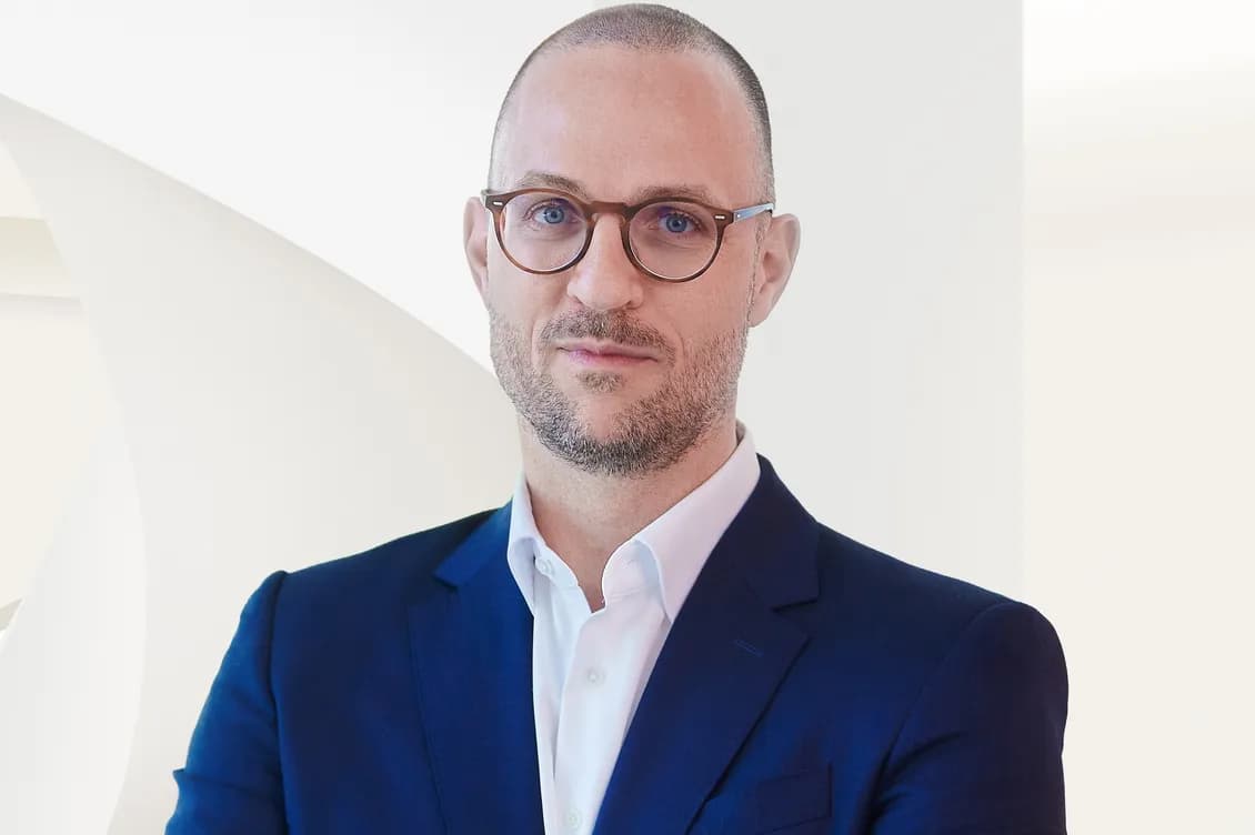 Bald man with round glasses and light stubble wearing a navy suit and white shirt against a pale geometric background.