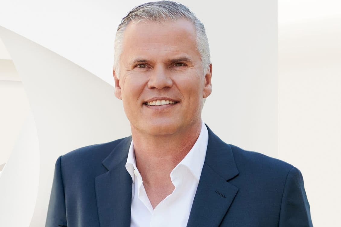 Smiling middle-aged man with short gray hair in a dark suit and open-collar white shirt against a pale geometric background.
