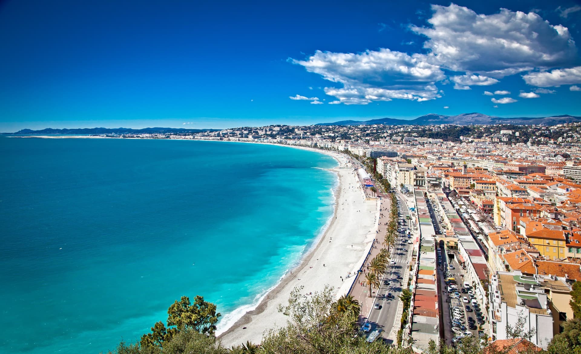 Scenic view of Nice, France, featuring turquoise sea, white beach, and buildings under a blue sky with scattered clouds.
