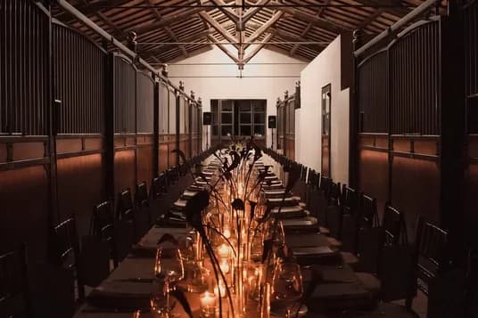 Dimly lit dining hall with exposed wooden beams; a long banquet table lined with candles, glassware, and place settings on both sides.