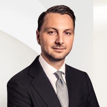 Frederic Weber in einem dunklen Anzug und einer hellen Krawatte posiert selbstbewusst vor einem hellen, abstrakten Hintergrund.